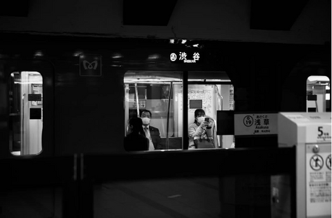 People inside a Tokyo train, seated or standing, with some reading or using their phones, and the train's interior visible, including overhead handles and seat designs.